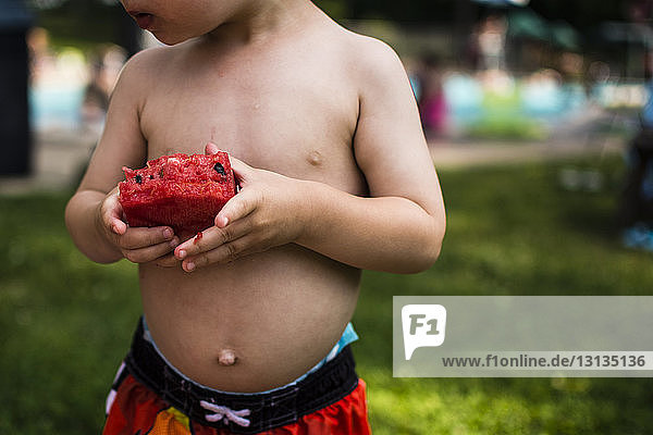 Midsection of shirtless boy eating watermelon while standing in backyard