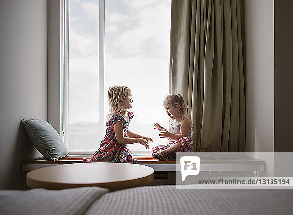 Happy sisters talking while sitting on table by window at home