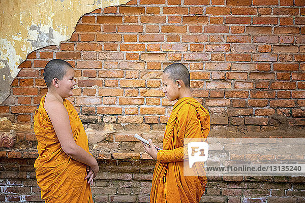 Seitenansicht von buddhistischen Novizenmönchen  die im Stehen an einer Ziegelmauer studieren