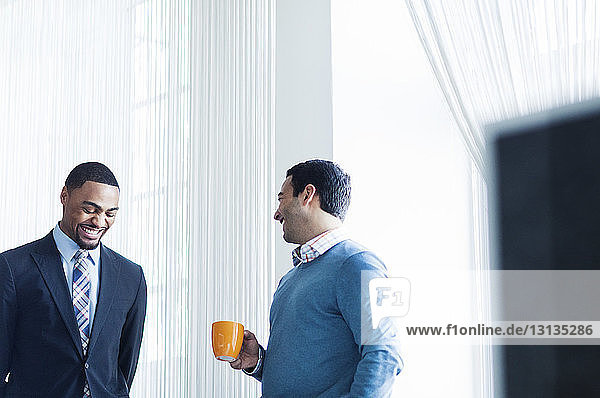 Geschäftsmann hält Kaffeetasse in der Hand  während er sich mit einem glücklichen Kollegen unterhält