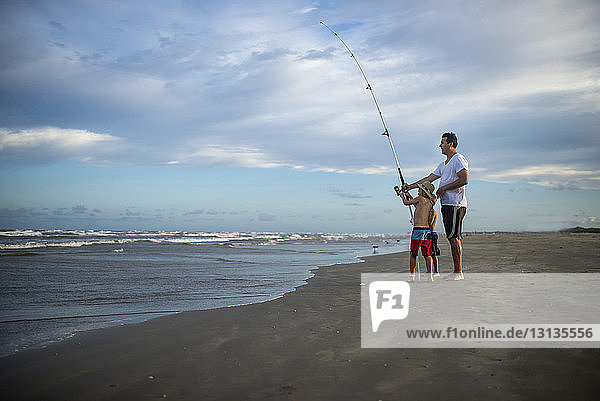 Verspielter Vater und Söhne mit Angelrute stehen am Strand vor wolkigem Himmel