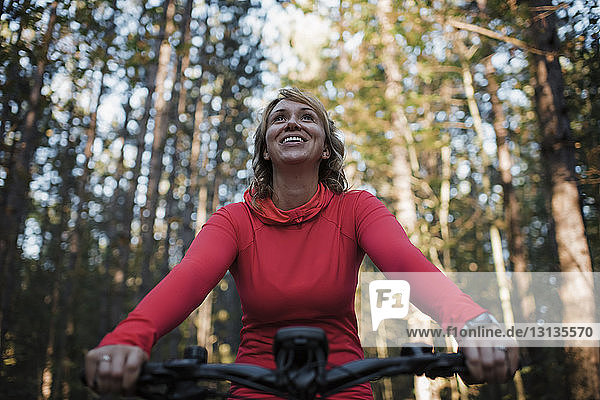 Low angle view of cheerful woman mountain biking against trees in forest