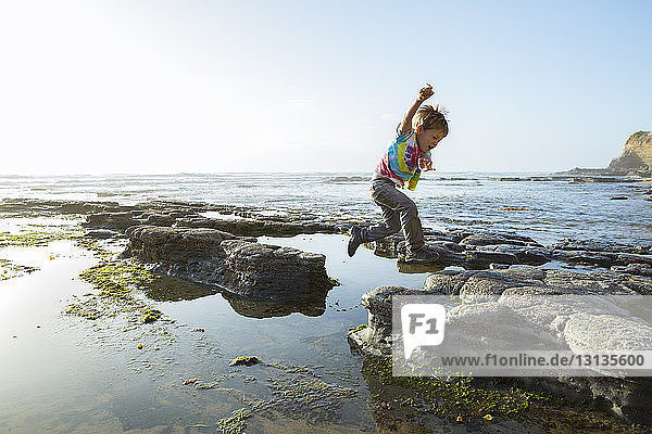 Side view of boy jumping on rocks at beach against clear sky