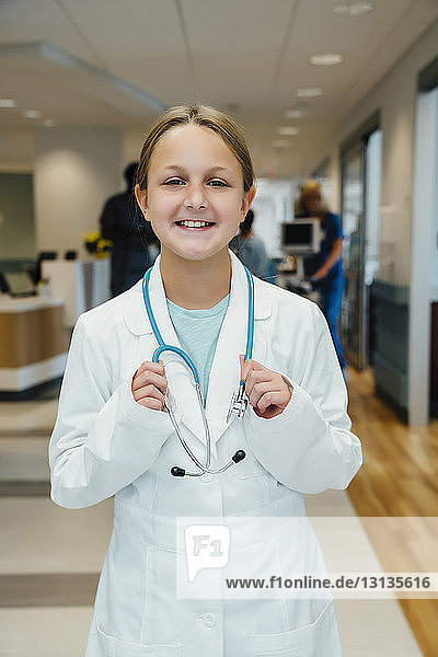 Portrait of confident girl with stethoscope wearing lab coat while standing in hospital corridor