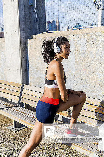Female athlete listening music while exercising on bench during sunny day