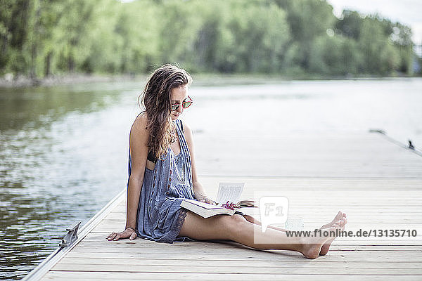 Woman reading book while resting on pier over lake