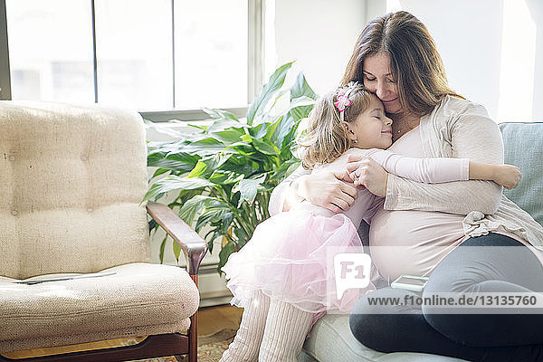 Happy pregnant woman embracing daughter while sitting on sofa at home