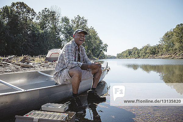 Happy man holding fishing rod while sitting on boat at lakeshore