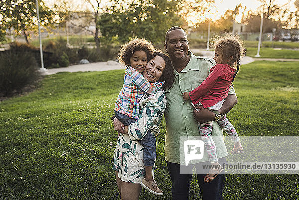 Porträt von fröhlichen Eltern  die ihre Kinder bei Sonnenuntergang auf dem Feld im Park tragen