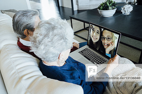 Senior couple video conferencing with granddaughters through laptop computer while sitting on sofa at home