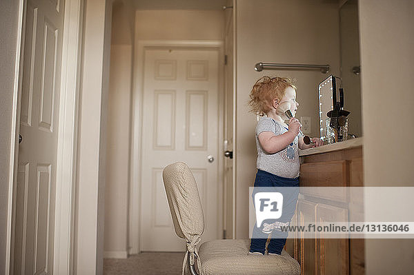 Cute baby boy applying make-up while standing on chair at home