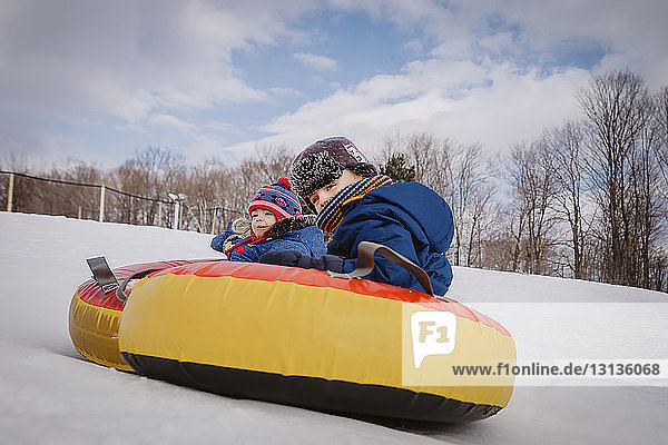 Siblings tobogganing with tube sled on snow covered hill against cloudy sky