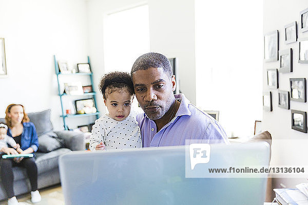 Tochter und Vater schauen auf Laptop-Computer  während Mutter und Schwester im Hintergrund sitzen