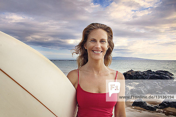 Porträt einer lächelnden Frau mit Surfbrett am Meeresufer vor bewölktem Himmel