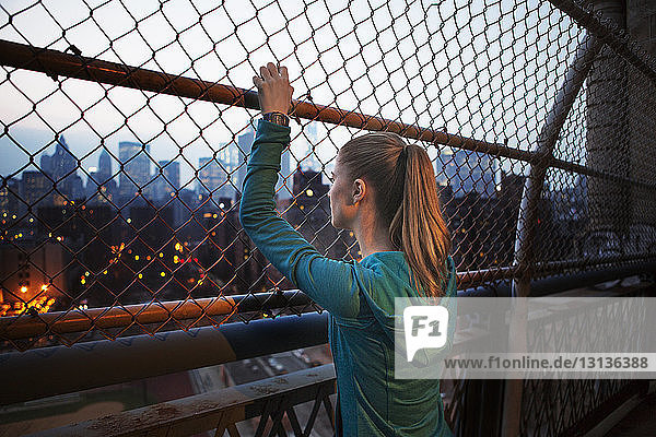 Nachdenkliche junge Frau schaut durch den Maschendrahtzaun  während sie auf einer Brücke in der Stadt steht