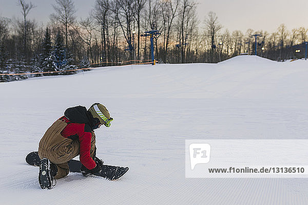Full length of teenage boy strapping snowboard on snow covered landscape during sunset