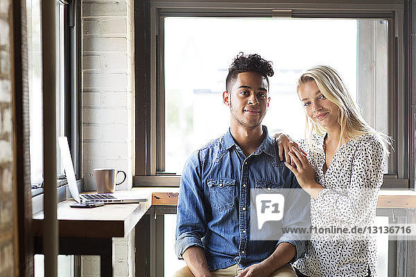 Portrait of smiling couple by laptop against windows at cafe