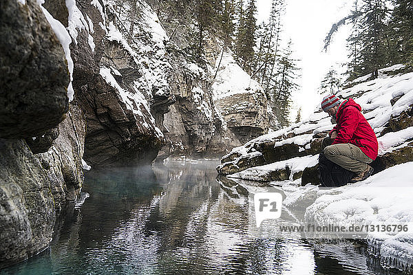 Side view of hiker with backpack crouching by stream at forest during winter