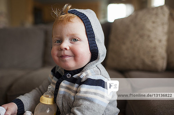 Portrait of cute baby boy with milk bottle wearing warm clothing in living room at home