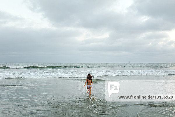 Rear view of shirtless girl running towards sea against cloudy sky at beach