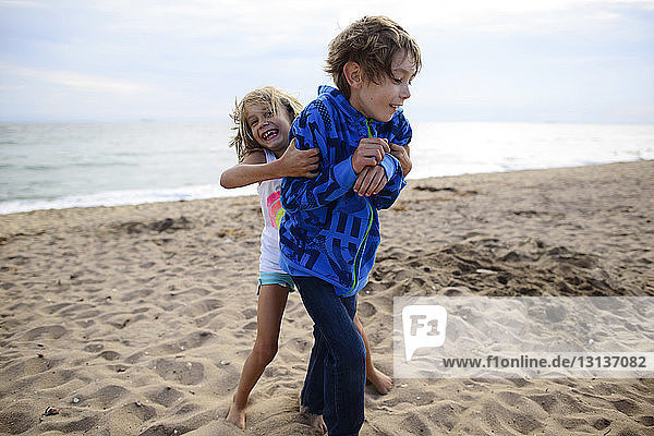 Playful sister tickling brother while standing at Huntington Beach against sky