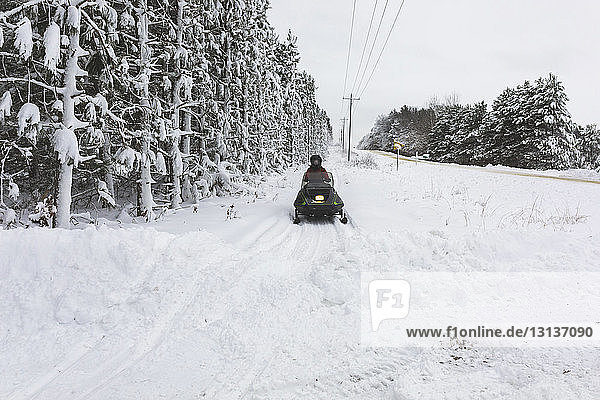 Rear view of teenage boy driving quadbike on snow covered field against sky in forest