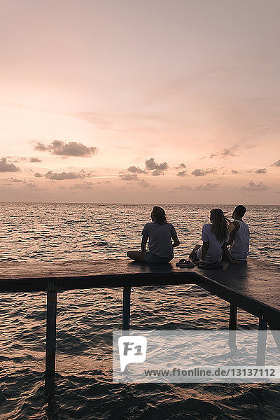 Scherenschnitt-Freunde sitzen bei Sonnenuntergang am Pier über dem Meer gegen den Himmel