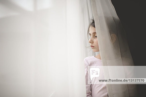 Mädchen mit geschlossenen Augen steht zu Hause am Fenstervorhang