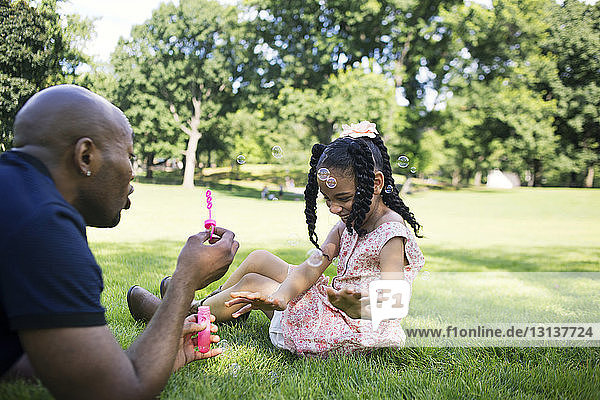 Vater und Tochter spielen mit Seifenblasen auf Grasfeld im Park