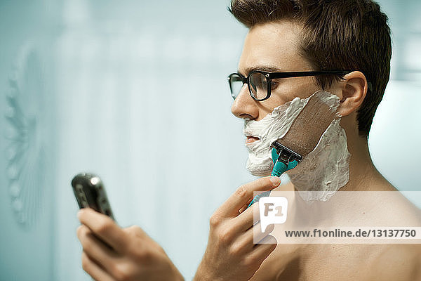 Mann benutzt Telefon beim Rasieren des Gesichts zu Hause