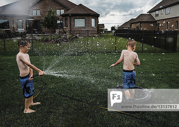 Junge ohne Hemd spritzt mit Gartenschlauch Wasser auf Bruder im Park