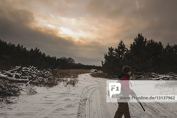 Side view of boy with rifle walking on snowy field against cloudy sky