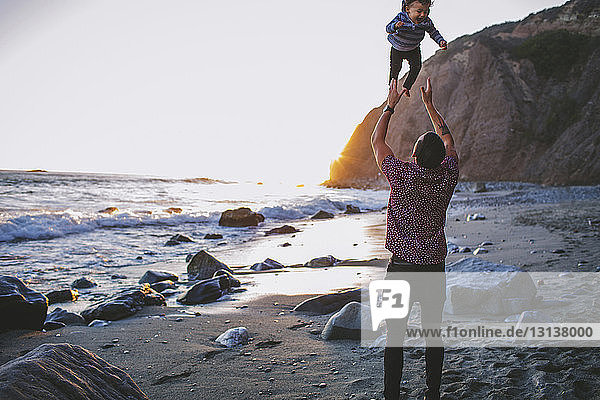 Rückansicht eines spielerischen Vaters  der seinen Sohn am Strand in die Luft wirft
