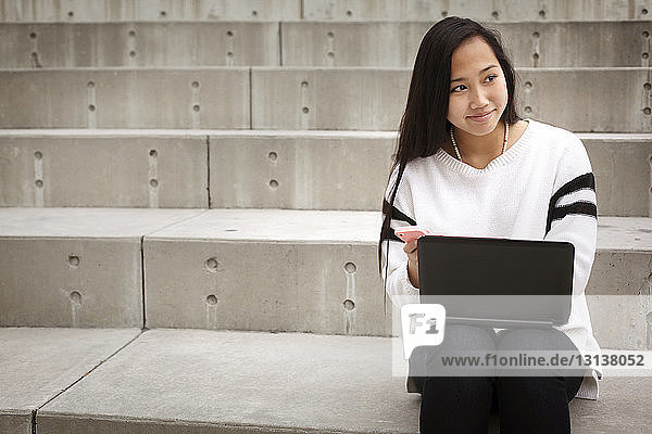 Teenager looking away while sitting on steps with laptop