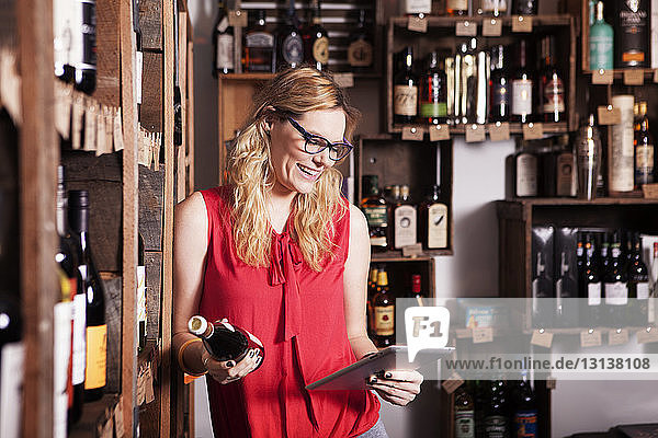 Glückliche Unternehmerin benutzt Tablet-Computer  während sie eine Weinflasche im Laden hält
