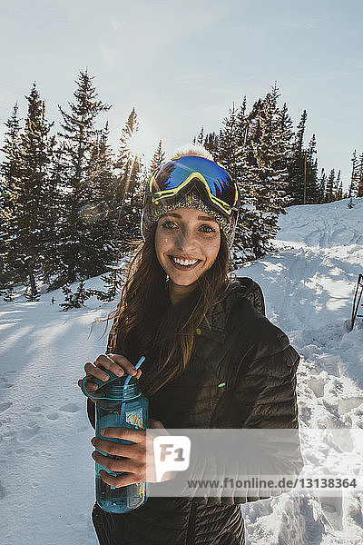 Portrait of smiling young woman wearing ski goggles while holding water bottle on snowy field