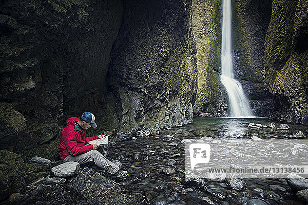 Mann liest Karte  während er am Wasserfall im Wald sitzt