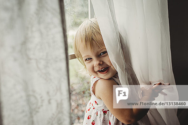 Porträt eines fröhlichen Mädchens  das zu Hause am Fenster mit Vorhängen spielt