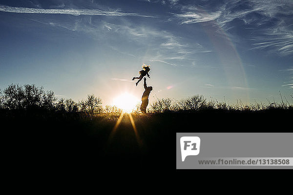 Verspielter Silhouettenvater  der seine Tochter in die Luft wirft  während er mit ihr bei Sonnenuntergang auf dem Feld gegen den Himmel spielt