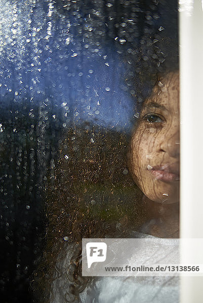 Mädchen schaut weg  während sie zu Hause steht und durch nasses Fensterglas gesehen wird