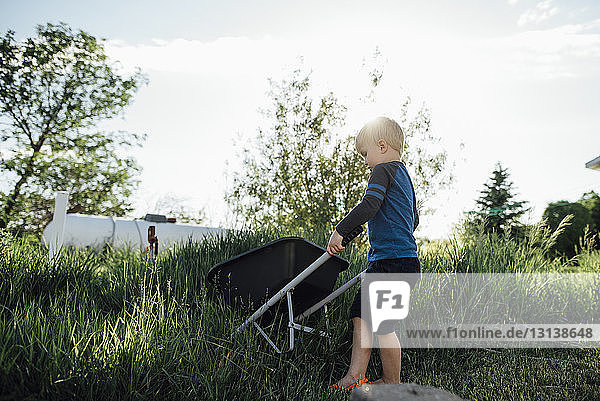Side view of boy holding wheelbarrow while standing on grassy field against sky