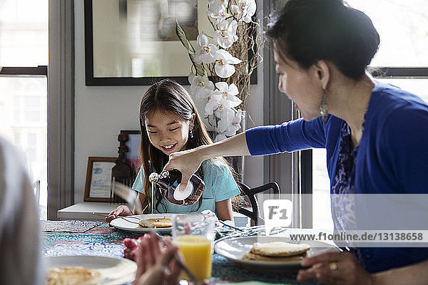 Mother pouring maple syrup on pancakes for daughter during breakfast