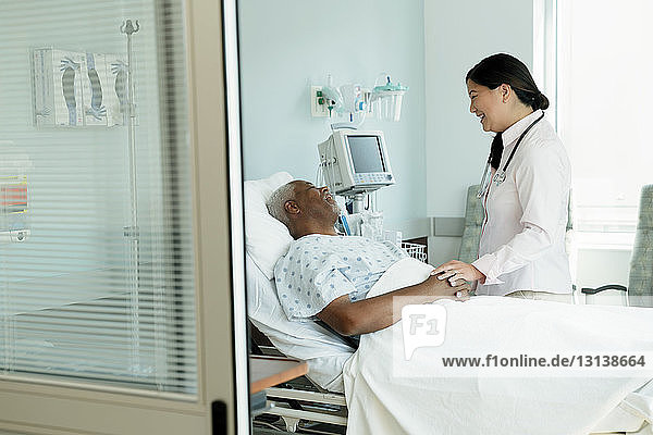 Cheerful doctor talking to senior patient lying on bed in hospital ward