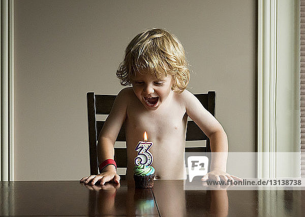 Fröhlicher Junge mit offenem Mund schaut Geburtstagskuchen an  während er am Tisch steht