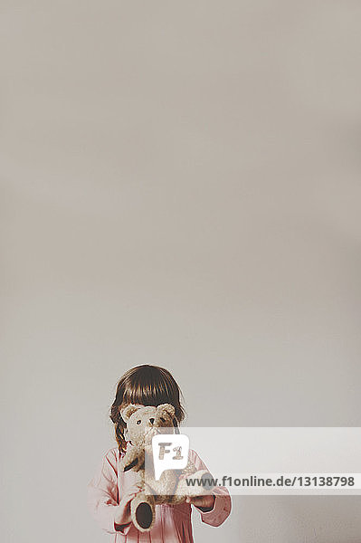 Girl holding teddy bear in front of face against white wall