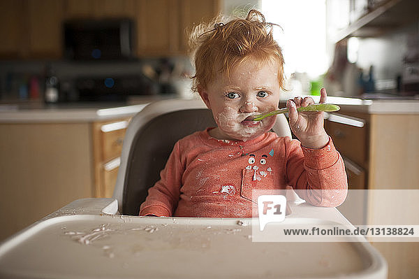 Porträt eines süßen kleinen Jungen mit unordentlichem Gesicht  der zu Hause auf einem Hochstuhl sitzend Essen isst