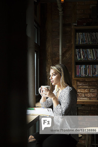 Frau mit Zeitung  die Kaffee trinkt  während sie durch ein Fenster auf ein Café schaut