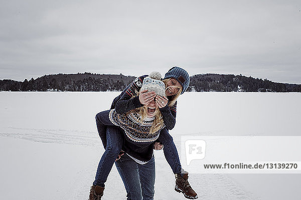 Verspielte Frau  die die Augen ihrer Freundin bedeckt  während sie im Winter von ihr auf Schnee huckepack getragen wird
