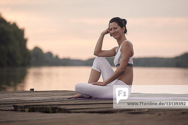 Porträt einer Frau  die Yoga praktiziert  während sie bei Sonnenuntergang auf einem Pier am See vor bewölktem Himmel sitzt