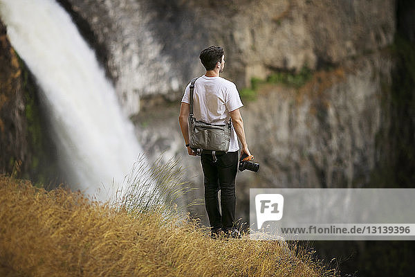 Rear view of hiker holding camera while standing on mountain against waterfall